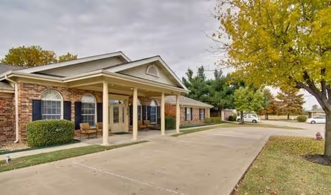 Exterior view of a single-story brick building with a covered entrance supported by columns, surrounded by trees with autumn foliage and a paved driveway in front.