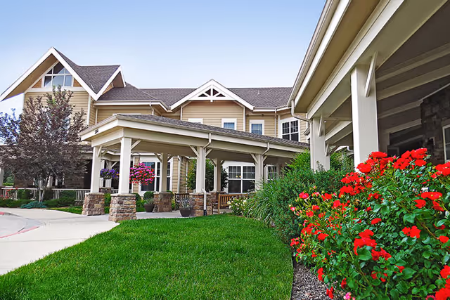 Exterior view of MorningStar Assisted Living & Memory Care of Littleton showing a covered entrance with stone pillars, beige siding, multiple windows, a well-maintained lawn, and vibrant red flowers in the foreground.