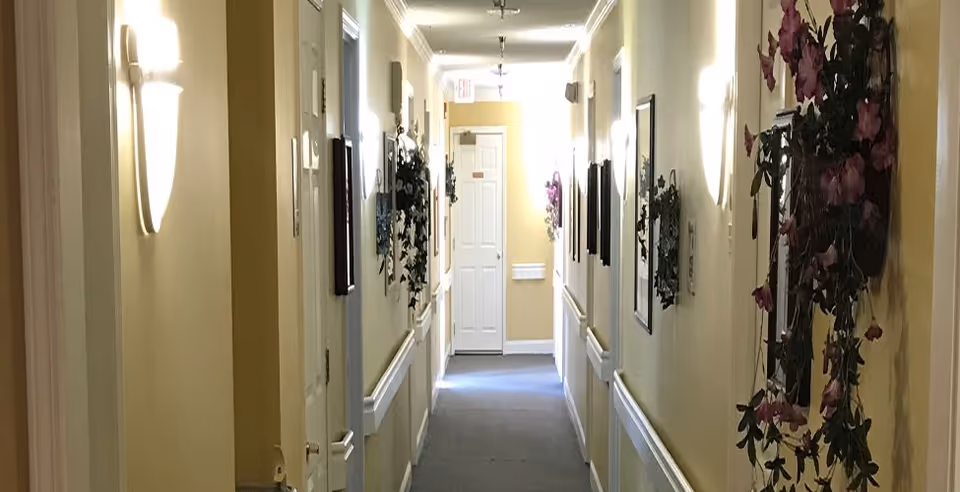 A well-lit indoor hallway with beige walls and a gray carpet. The hallway is decorated with wall-mounted light fixtures, framed pictures, and hanging flower arrangements. Several doors line the hallway, and there is an exit sign above a door at the far end.