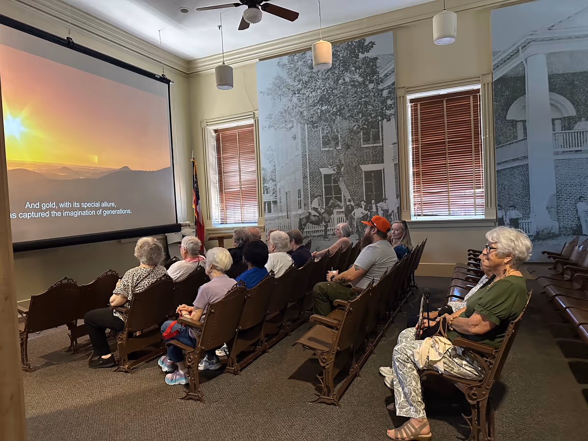 A group of elderly people seated in rows of vintage theater-style chairs watching a presentation or movie projected on a screen in a room with large black and white historical photos on the walls and windows with wooden blinds.
