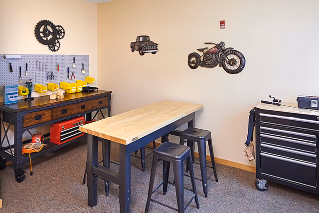 A workshop room with a wooden workbench and three metal stools. On the left side, there is a pegboard with various tools hanging and yellow bins on a wooden and metal table. On the right side, there is a black tool chest on wheels. The walls are decorated with metal art of a motorcycle and a vintage truck.
