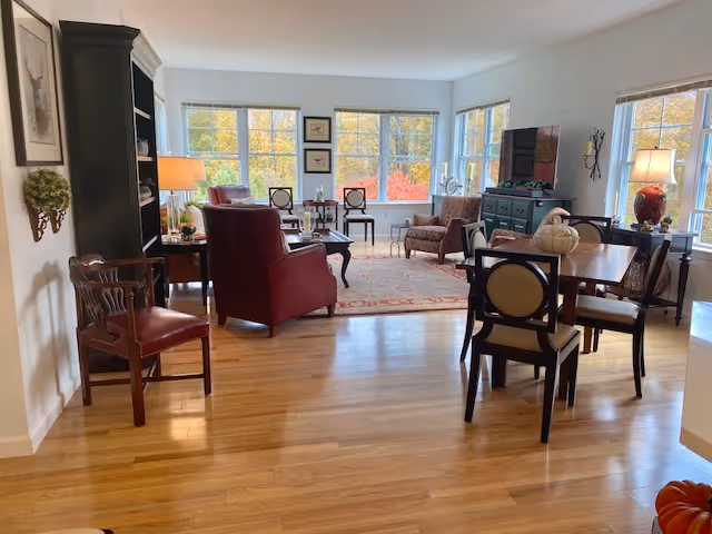A bright and spacious living room area with large windows showing autumn trees outside. The room features wooden flooring, a red armchair, a wooden dining table with chairs, a dark green cabinet with a TV on top, and various lamps and framed artwork on the walls.