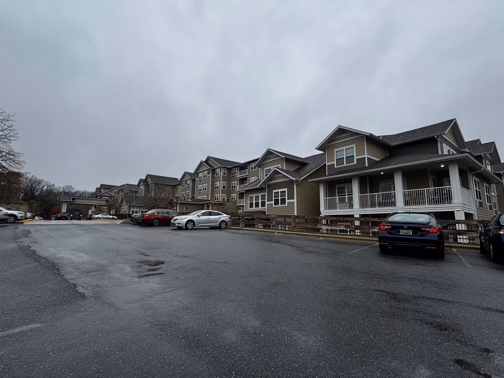 Exterior view of a multi-story residential building with beige siding and white trim under a cloudy sky. Several cars are parked in an asphalt parking lot in front of the building.