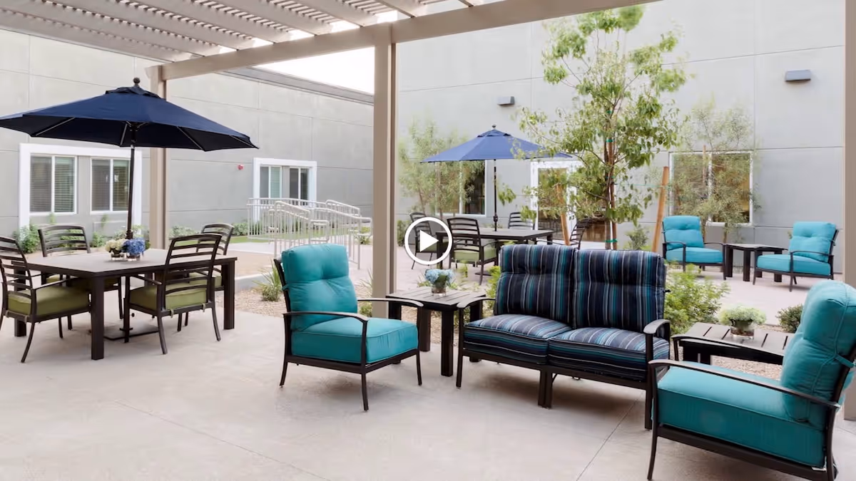 Outdoor patio area with cushioned seating including armchairs and a loveseat, tables with flower arrangements, and dining tables with umbrellas under a pergola. The space is surrounded by a building with windows and some greenery.