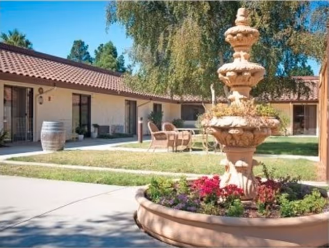 Outdoor courtyard area with a multi-tiered stone fountain surrounded by colorful flowers. There are patio chairs and tables on a grassy area, with single-story buildings featuring tiled roofs and large windows in the background under a clear blue sky.