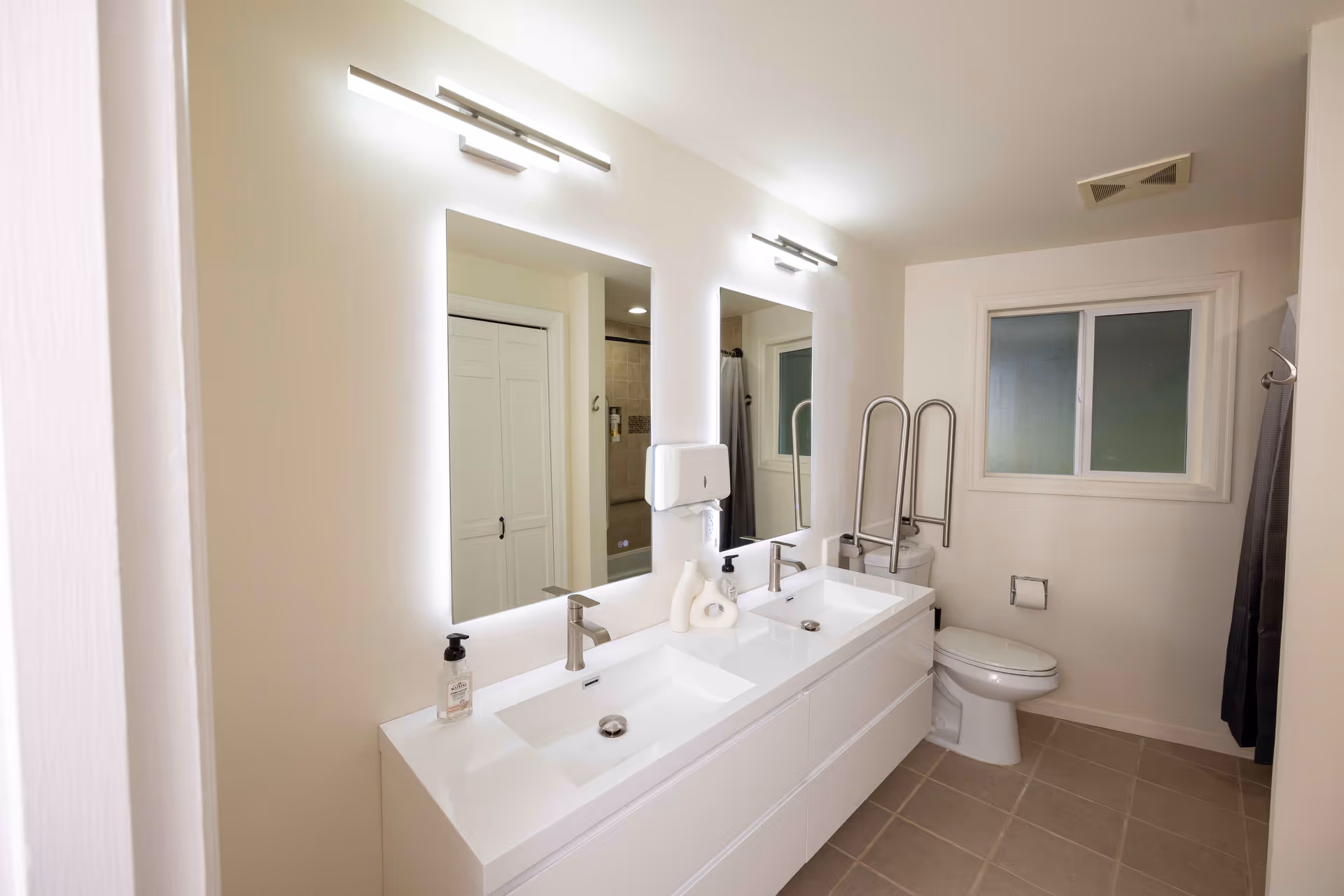 A clean, modern bathroom with a double sink vanity featuring two illuminated mirrors above. There is a white toilet with safety grab bars next to it, a window with frosted glass, and a tiled floor. The walls are painted white, and there is a soap dispenser and lotion bottles on the vanity.