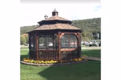 A wooden octagonal gazebo on a grassy lawn with flowerbeds, parked cars, and tree-covered hills in the background.