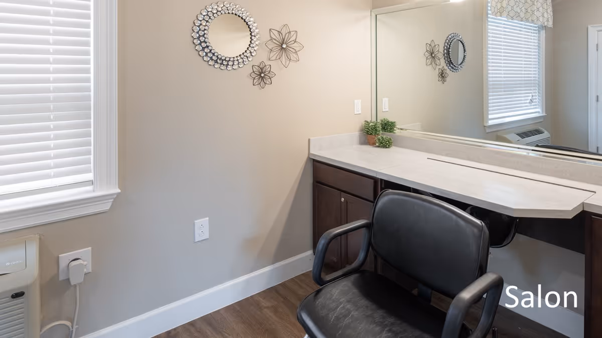 Interior view of a salon area with a black salon chair in front of a countertop and large mirror. The wall has decorative round and flower-shaped metal wall art. A window with white blinds is on the left side, and there is a small potted plant on the countertop.