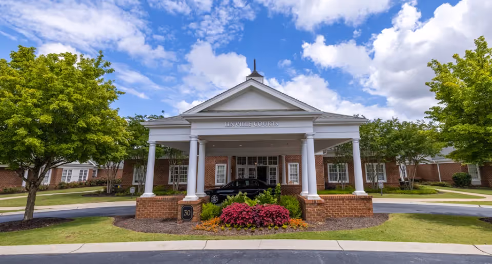Front exterior view of a senior living facility named Linville Courts with a covered entrance supported by white columns, surrounded by green trees and landscaped flower beds under a partly cloudy sky.