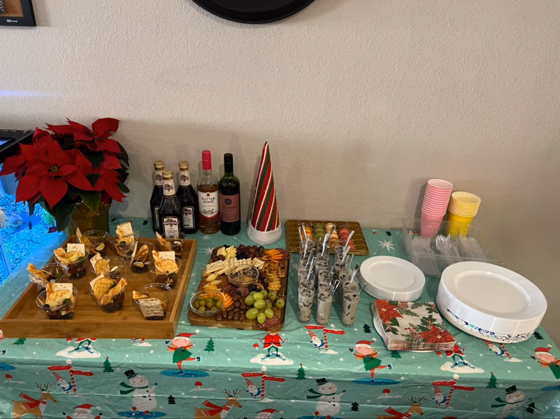 A festive table setup with a holiday-themed tablecloth featuring snowmen, elves, and candy canes. On the table are various snacks including crackers, grapes, olives, and small dessert cups. There are also bottles of wine and liquor, a red and green striped cone decoration, stacks of pink and yellow cups, plastic utensils, paper plates, and holiday-themed napkins. A red poinsettia plant is placed on the left side of the table.