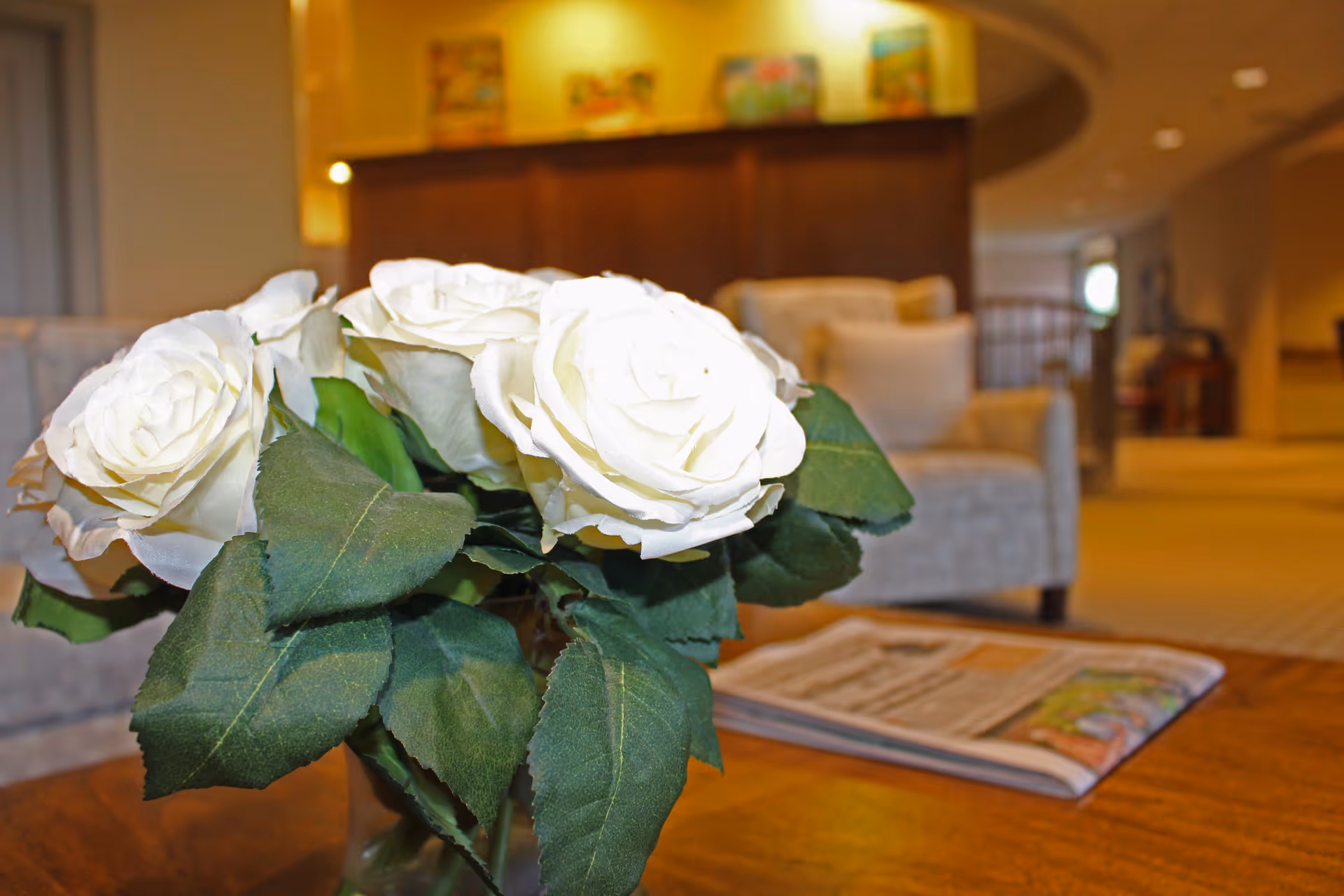 Close-up of a vase with white roses on a wooden table in a cozy living room area with beige armchairs and a newspaper on the table, soft warm lighting and blurred background.