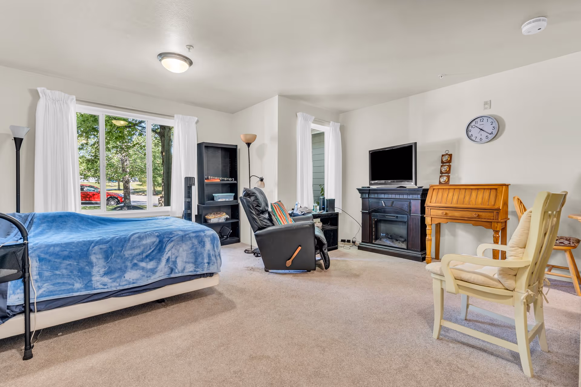 Bright studio-style bedroom and sitting area with a bed, recliner, TV above a fireplace, roll-top desk, and large windows showing trees outside.
