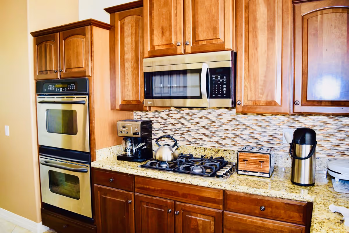 A kitchen area with wooden cabinets, a double oven, a microwave, a gas stove with a kettle on it, a coffee maker, a toaster, and a stainless steel airpot on a granite countertop. The backsplash features a mosaic tile design.