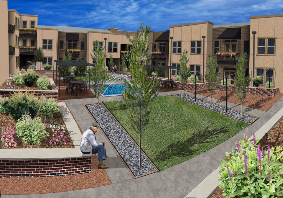 Outdoor courtyard area of a senior living facility with landscaped gardens, small trees, a water fountain, and seating areas. A person is sitting on a low brick wall. The building surrounds the courtyard with balconies and windows visible.