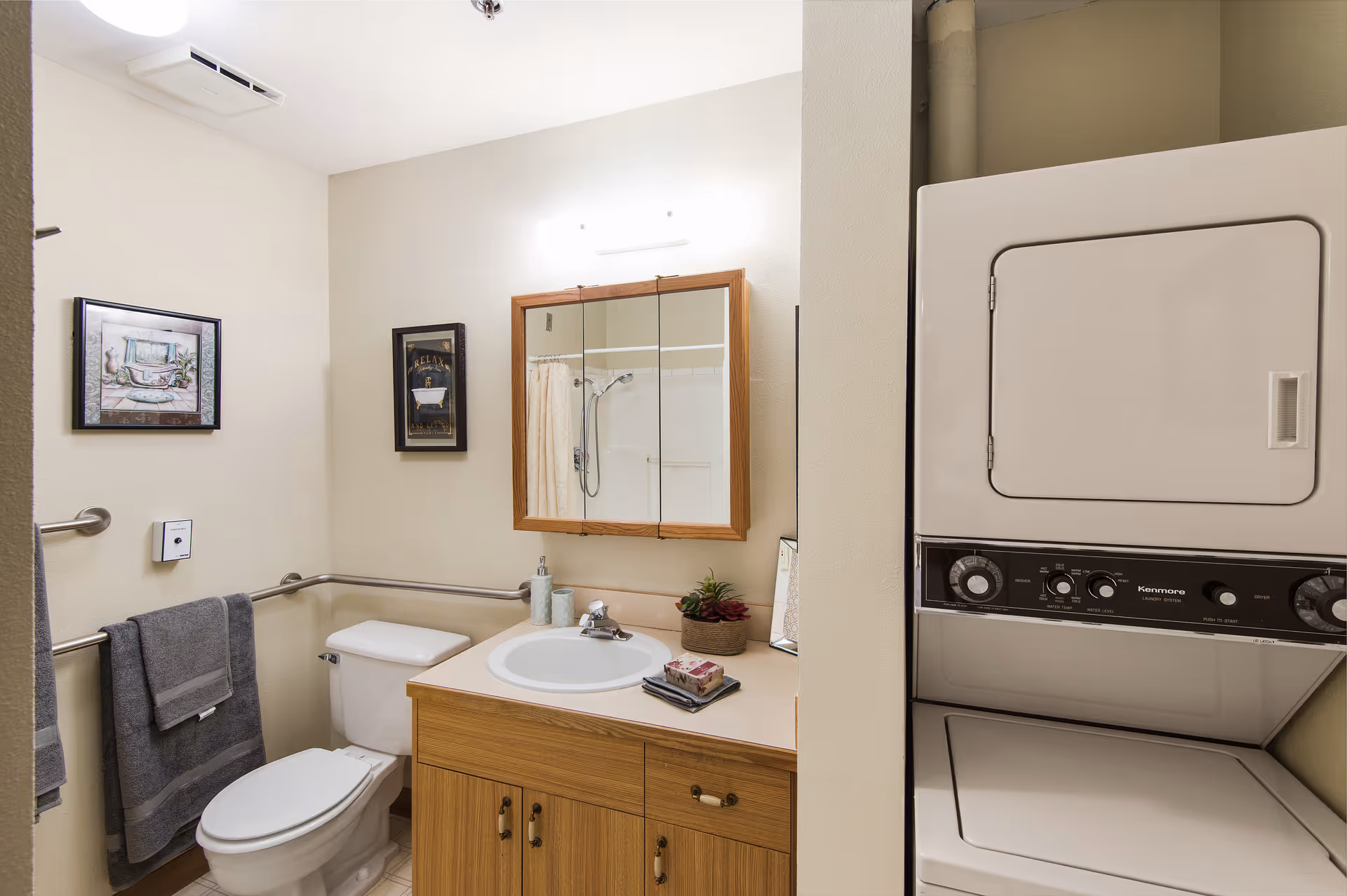 A small bathroom with a toilet, a sink with a wooden cabinet, a mirror cabinet above the sink, and a stacked washer and dryer unit to the right. The bathroom has beige walls, two framed pictures on the wall, and gray towels hanging on a metal bar.