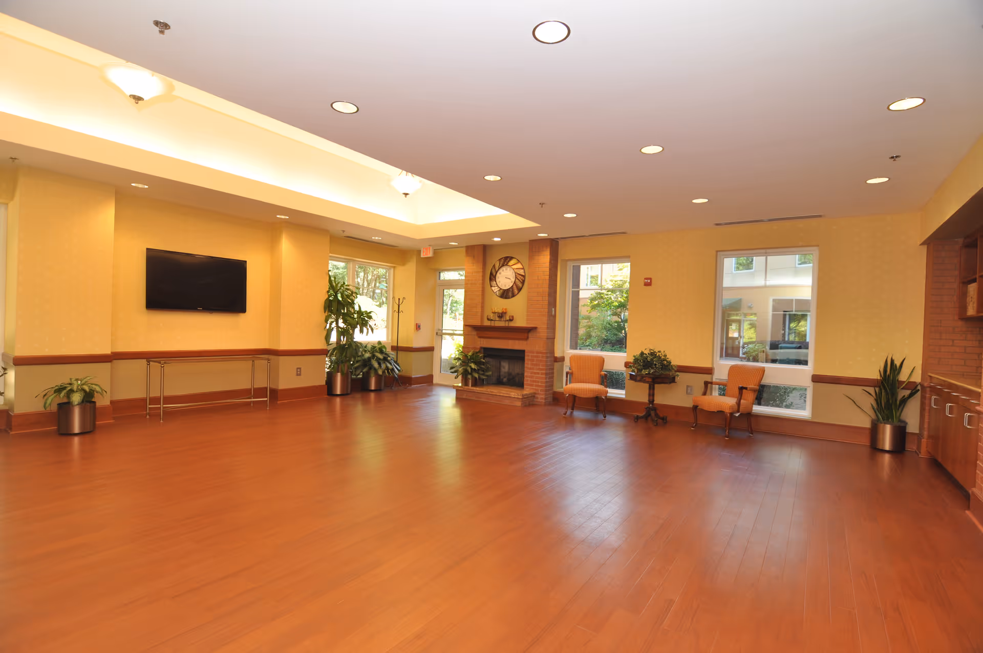 A spacious, well-lit common area with wooden flooring, yellow walls, and recessed ceiling lights. The room features a wall-mounted flat screen TV, several potted plants, a fireplace with a large clock above it, two orange upholstered chairs with a small table between them, and large windows letting in natural light.