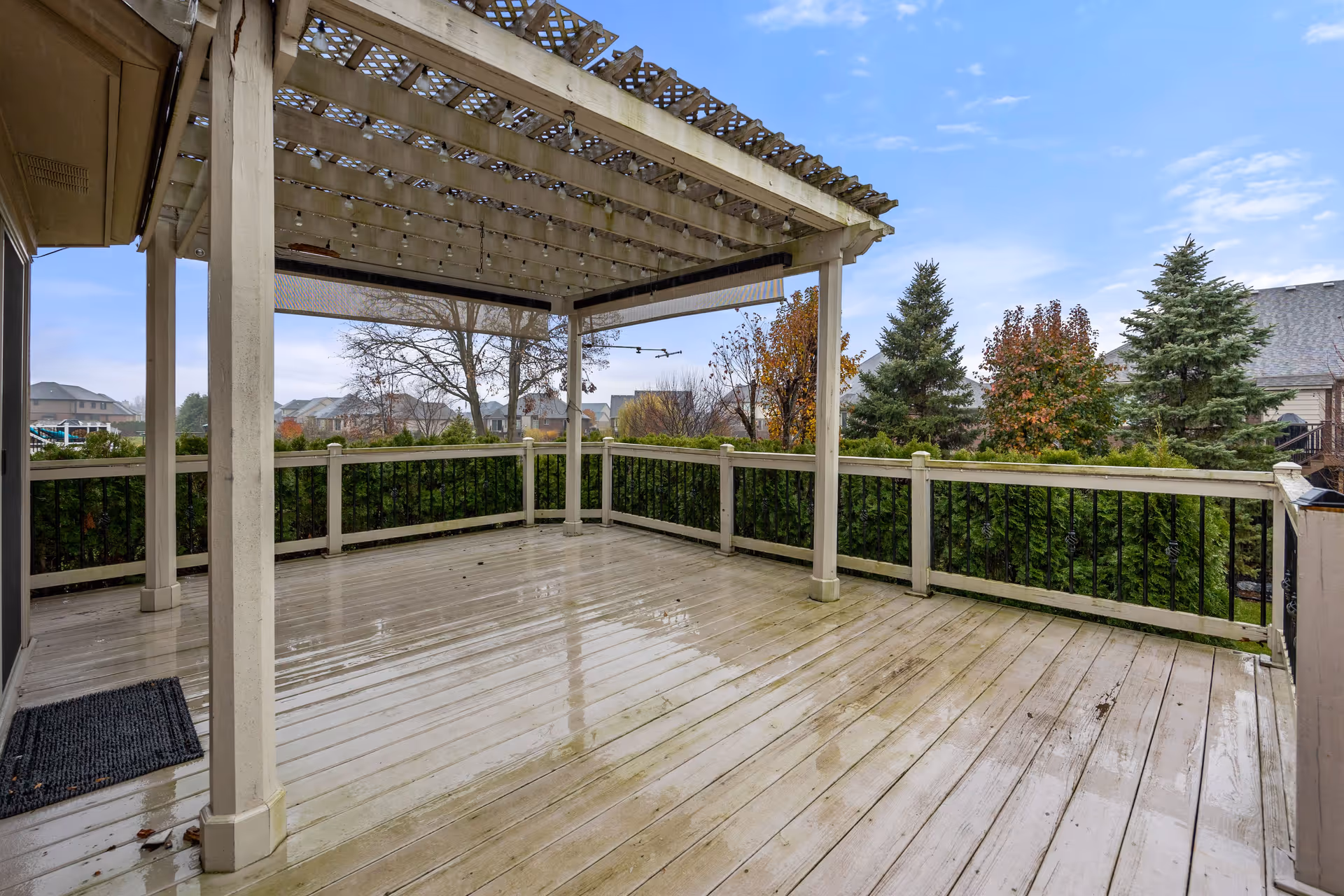 Wooden covered deck with a pergola and railing overlooking trees and neighboring houses.
