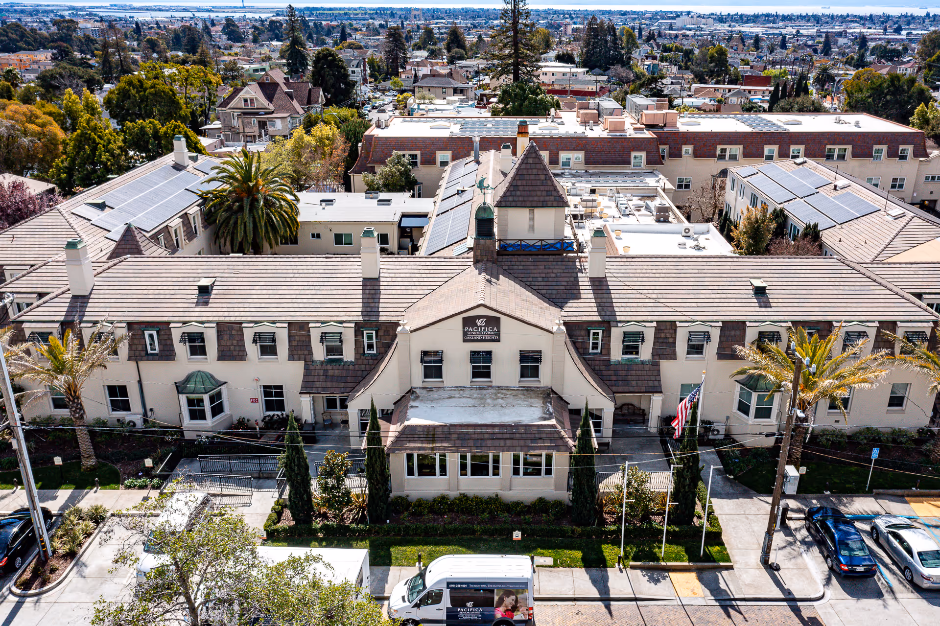 Aerial view of Oakland Heights Senior Living facility showing a large, multi-story building with a brown tiled roof and beige exterior. The building has multiple windows, a central entrance with a sign reading 'Pacifica Senior Living Oakland Heights,' and is surrounded by trees and parked vehicles, including a white van with the facility's branding. The background shows a residential neighborhood and distant cityscape.