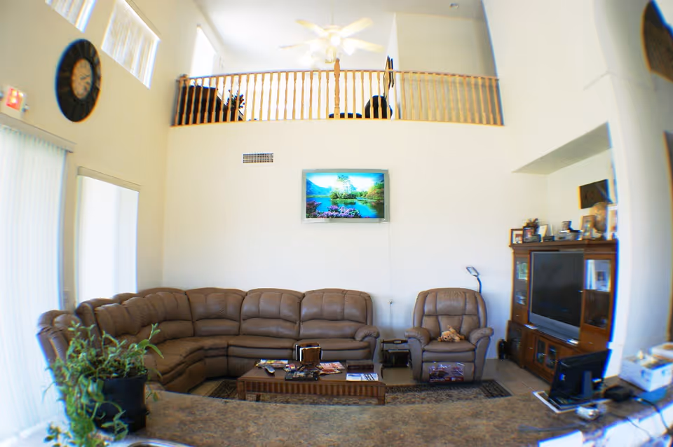 Bright two-story living room with a large brown sectional sofa, recliner, wall-mounted TV, and a wooden loft railing above.