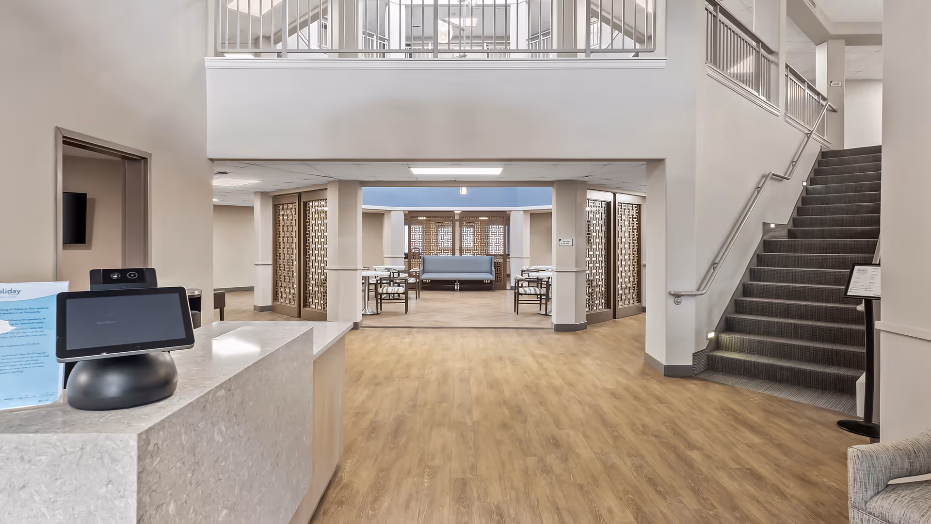 Interior view of a senior living facility lobby area with a reception desk on the left, a staircase with handrails on the right, and a seating area with tables and chairs in the background. The space features light-colored walls, wood flooring, and decorative partition panels.
