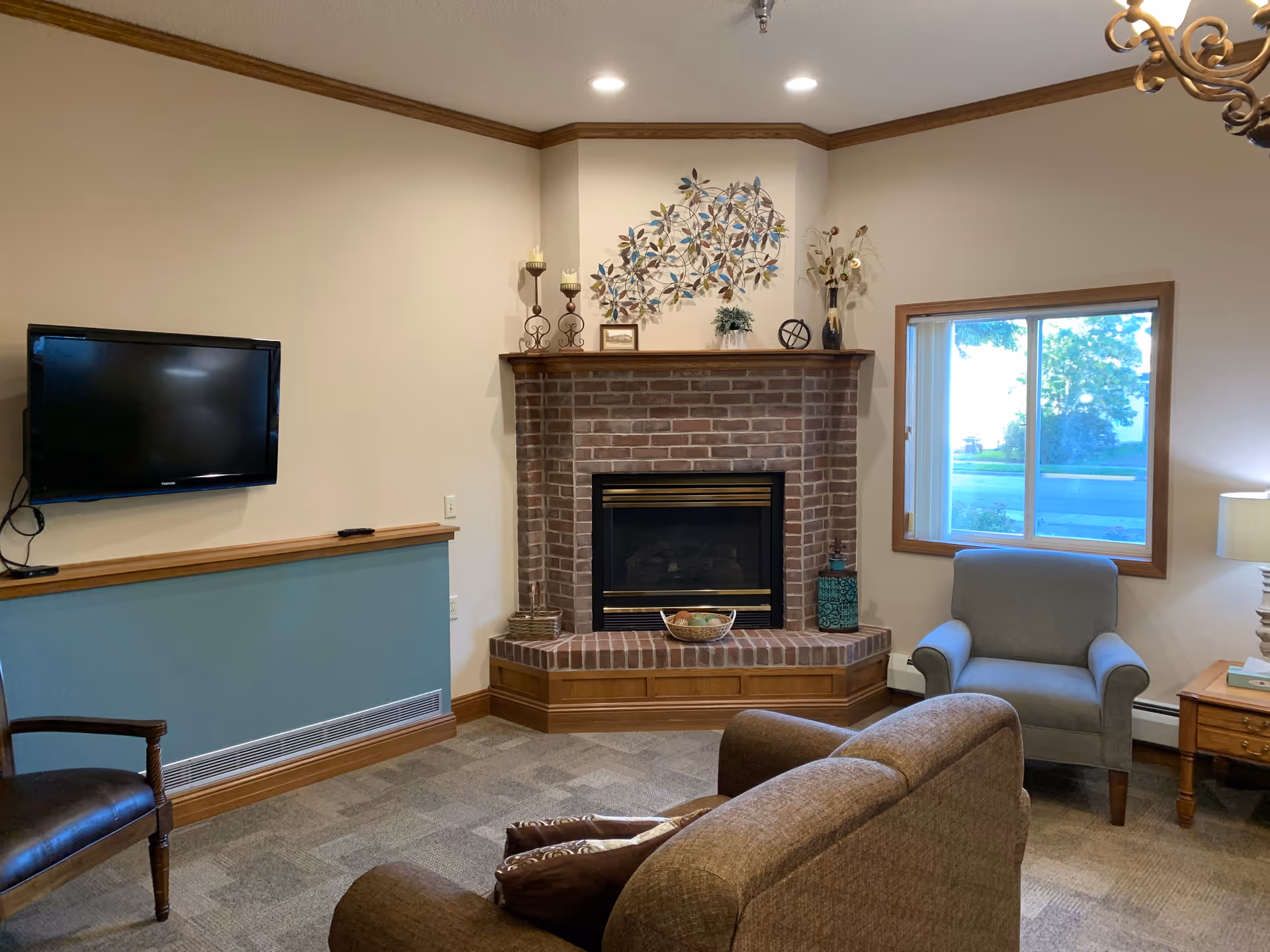 A cozy living room with a brick fireplace in the center, decorated with candles, a metal wall art piece, and a vase with flowers on the mantel. To the left, a flat-screen TV is mounted on the wall above a blue half-wall with wooden trim. There is a brown sofa facing the fireplace and a gray armchair near a window with a view outside. A wooden side table with a lamp and books is next to the armchair.