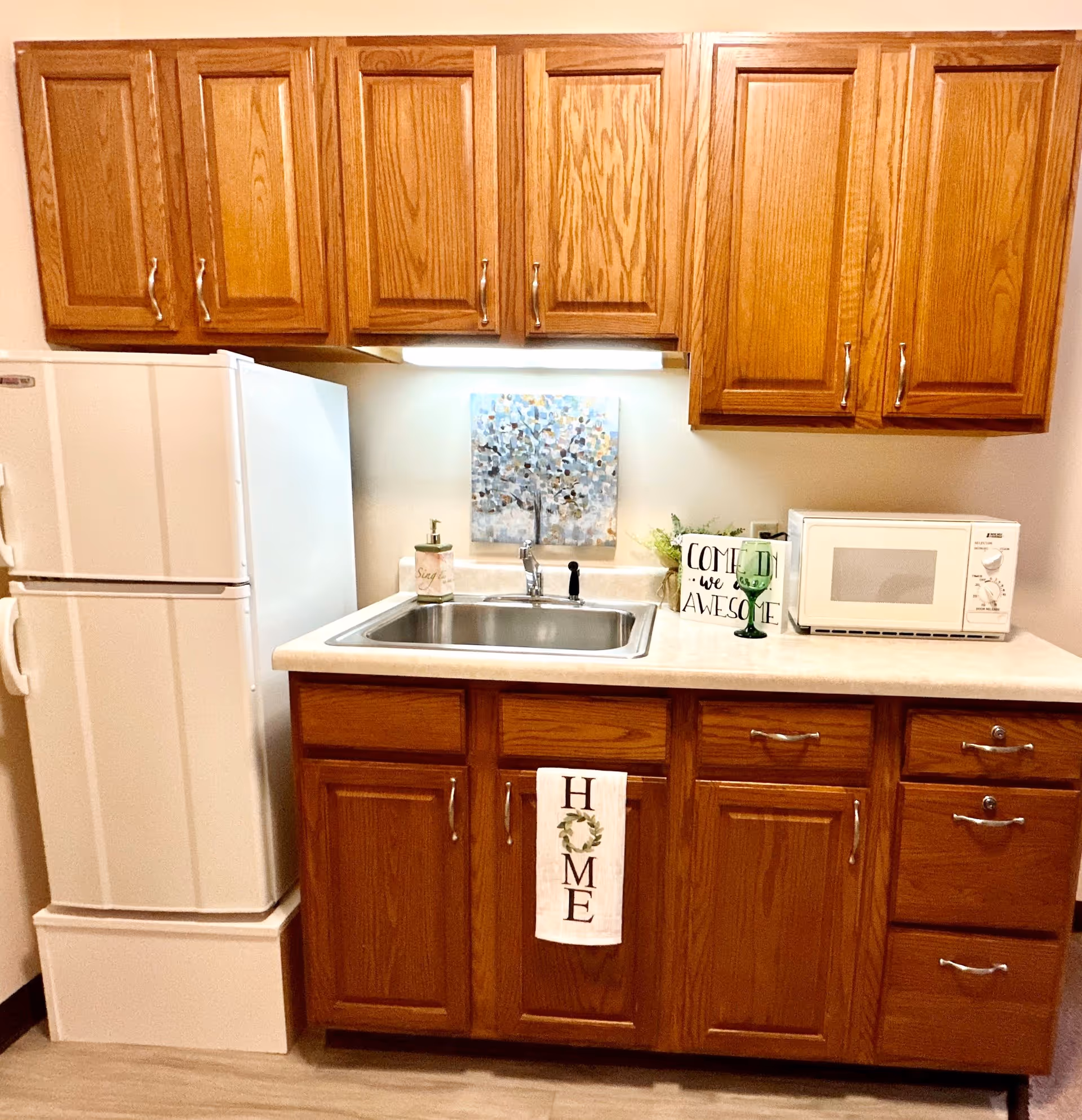 A small kitchen area with wooden cabinets above and below a countertop. The countertop has a stainless steel sink with a faucet, a soap dispenser, a green glass, a microwave, and a decorative sign that says 'COME IN we are AWESOME'. A white refrigerator is positioned to the left of the countertop. A white towel with the word 'HOME' hangs from the cabinet below the sink. There is a small painting of a tree on the wall above the sink.