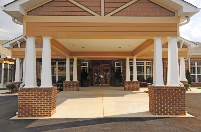 Front entrance of Webb House Retirement Center featuring a covered driveway supported by white columns with brick bases, double glass doors, and windows on either side.