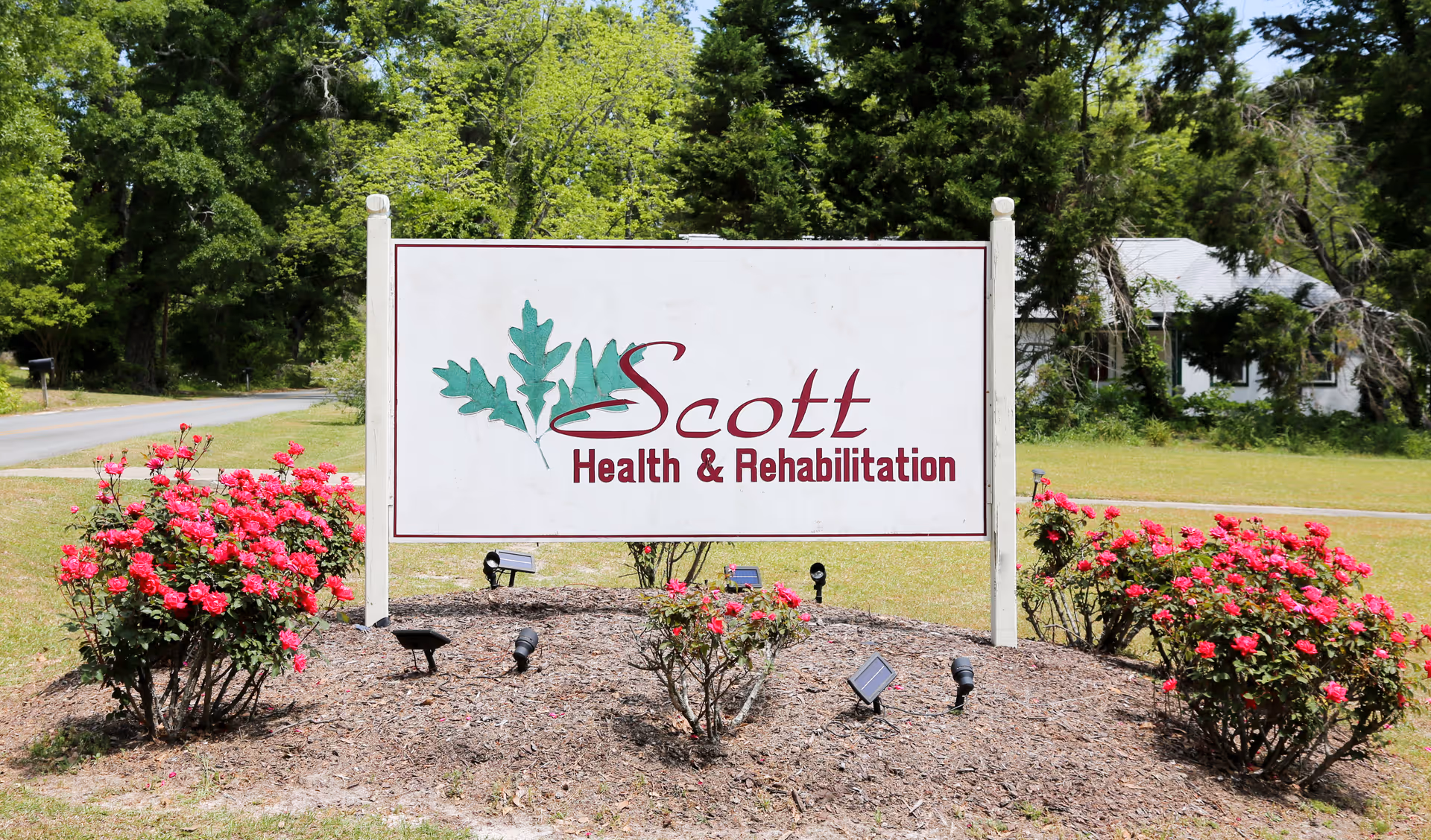 Outdoor sign for Scott Health & Rehabilitation surrounded by green trees and bushes with pink flowers, located near a road in a grassy area.