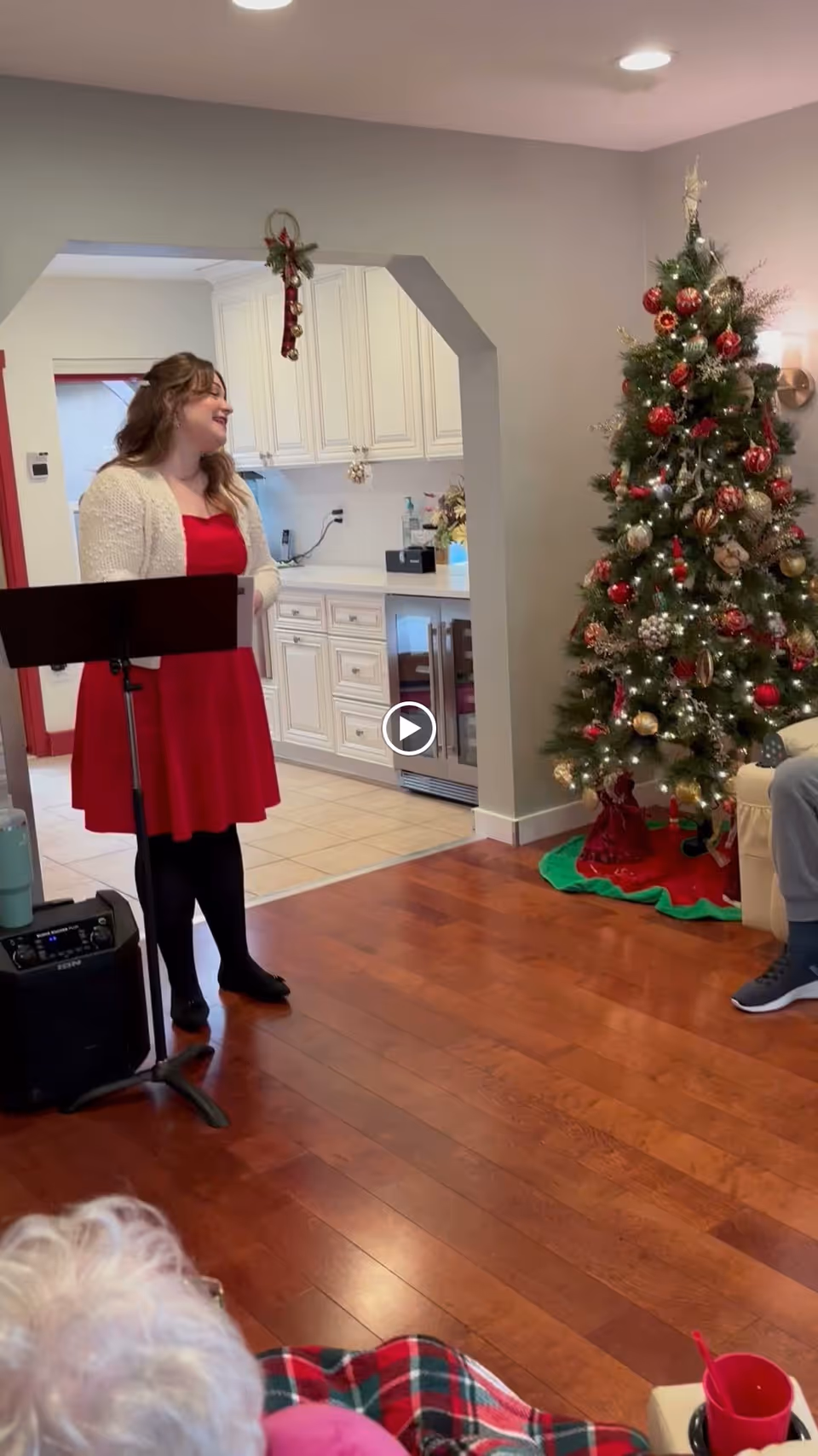 A woman in a red dress and white cardigan stands in a room with wooden floors, speaking or singing in front of a music stand. To her right is a decorated Christmas tree with red and gold ornaments. The room has white cabinetry and a small wine fridge in the background. An elderly person with white hair and a plaid blanket is partially visible in the foreground.