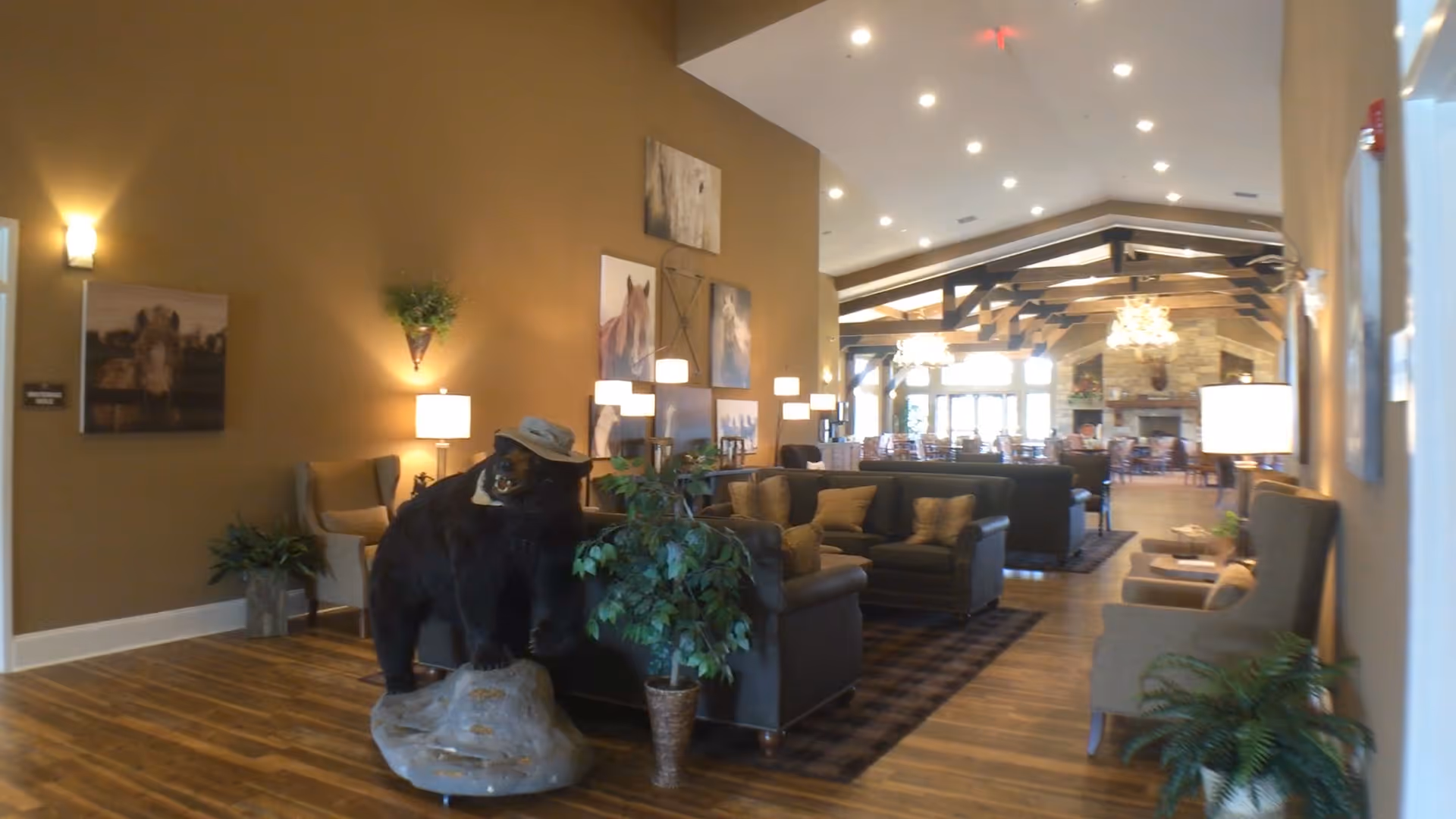 Interior view of a senior living facility lounge area with brown walls and wooden flooring. The room features comfortable seating including sofas and armchairs arranged around rugs. A decorative bear statue wearing a hat stands on a rock near a potted plant. The background shows a dining area with tables and chairs, exposed wooden beams on the ceiling, and a stone fireplace with chandeliers.
