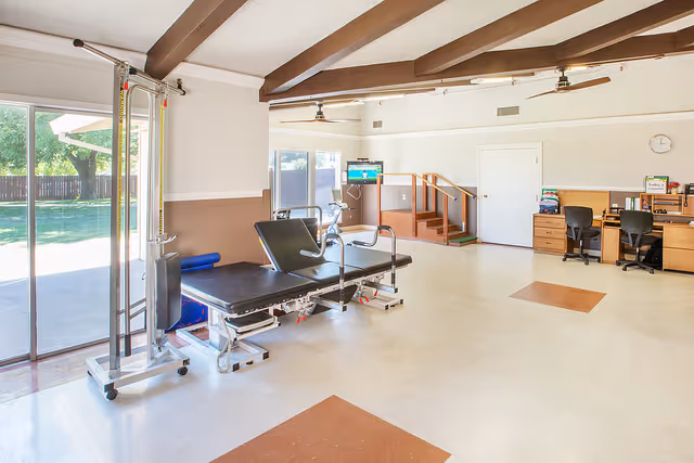 A spacious rehabilitation room with exercise equipment including a therapy table and pulley system. The room has large windows and a sliding glass door letting in natural light, a small set of stairs with a handrail for physical therapy, two desks with chairs and office supplies, and ceiling fans. The floor is tiled with some brown square accents.