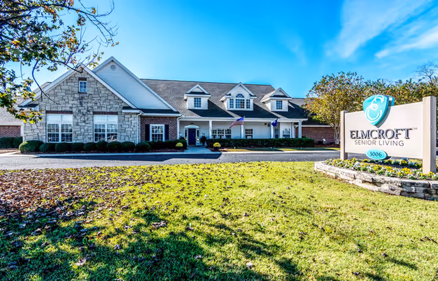 Front exterior of a single-story assisted living building with a lawn and a sign reading Elmcroft Senior Living.