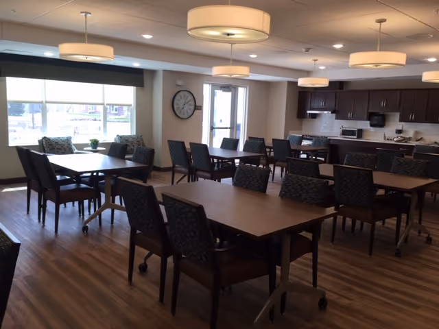 Well-lit dining area with multiple tables and chairs, pendant lights, large windows, and a kitchenette with cabinets and appliances.