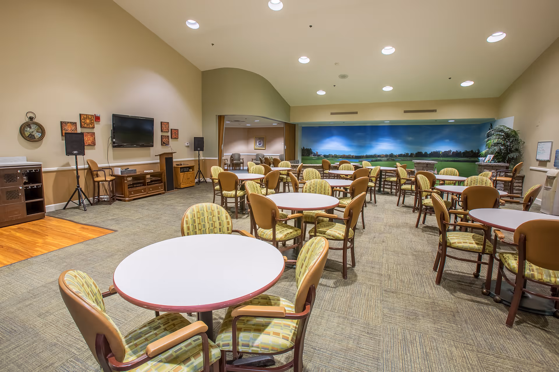 A spacious dining room with multiple round tables and cushioned chairs arranged neatly. The room has beige walls, a carpeted floor, and a large mural of a scenic outdoor landscape on the far wall. There is a TV mounted on one wall with speakers on stands nearby, and some decorative wall art around it.
