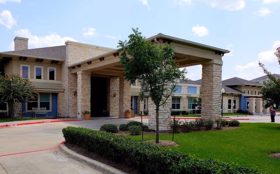 Front entrance of a senior living building with a stone porte-cochere, landscaped lawn and driveway.