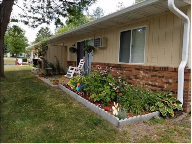 Single-story residential building with beige siding and brick lower half, featuring a small garden bed with flowers and plants along the side. A white rocking chair is placed near the entrance door, and there is a grassy lawn with trees nearby.