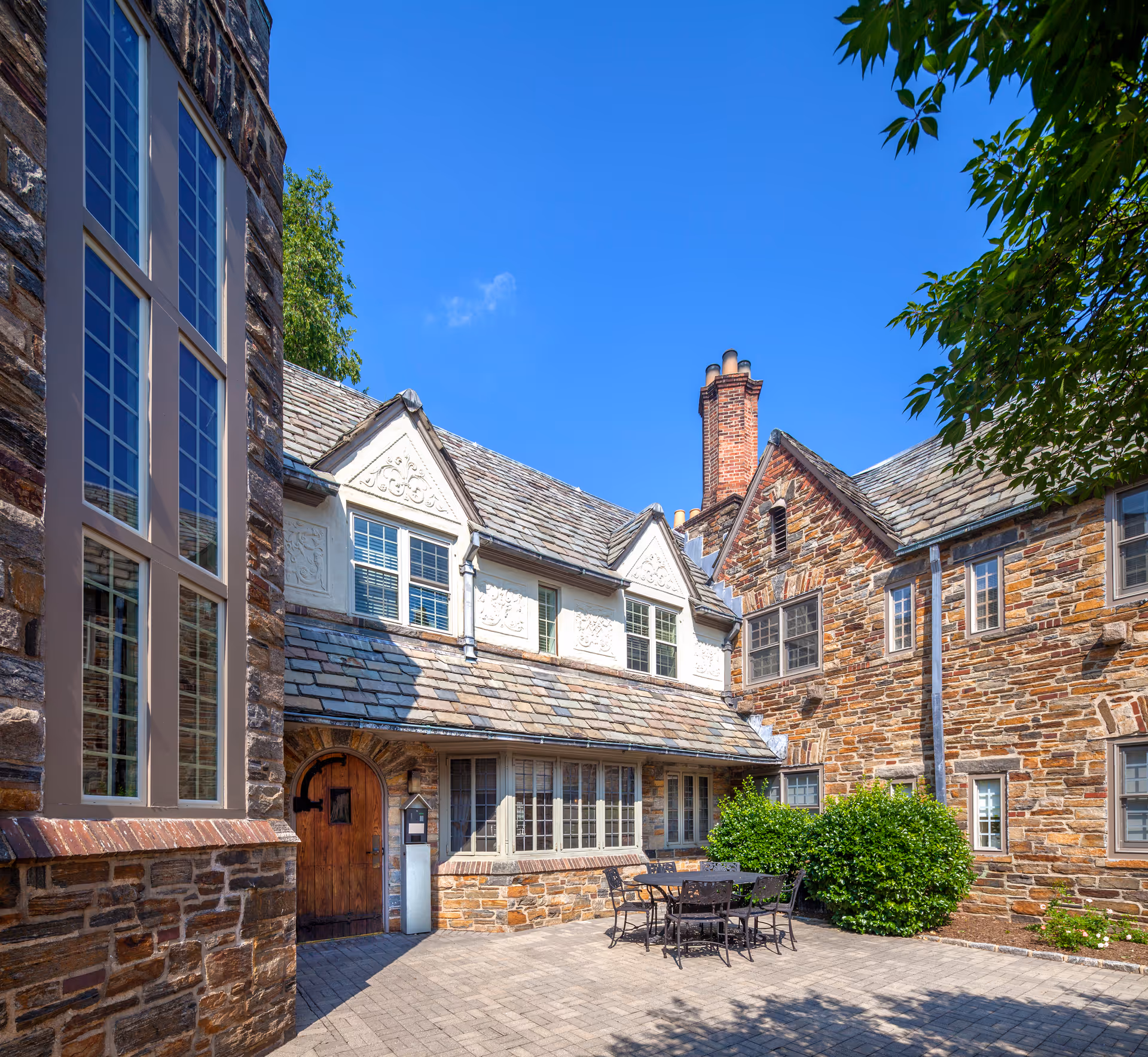 Outdoor courtyard area of a senior living facility with stone buildings, large windows, a wooden door, patio furniture including a table and chairs, and green bushes under a clear blue sky.