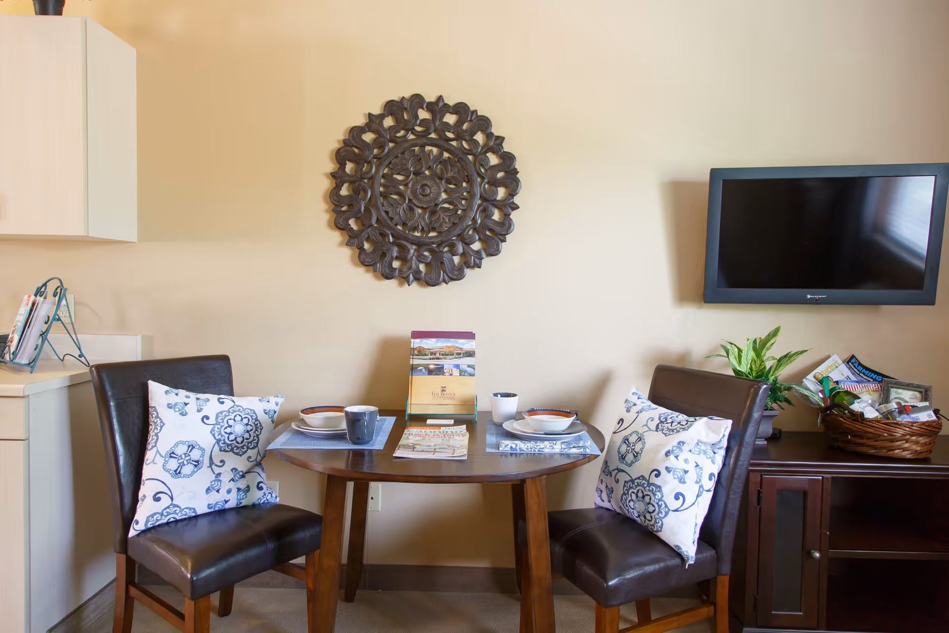 Small dining nook with a round wooden table set for two, two upholstered chairs with patterned pillows, wall-mounted TV, decorative wall art, and a side cabinet with a plant and basket.