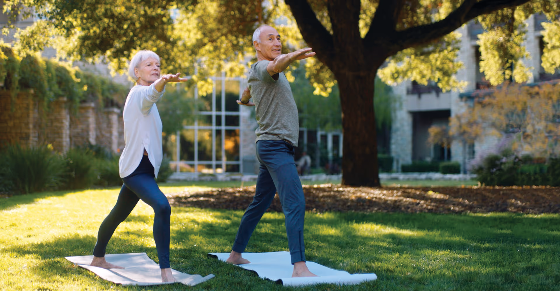 Two elderly individuals practicing yoga on mats outdoors in a grassy area with trees and buildings in the background, enjoying a sunny day.
