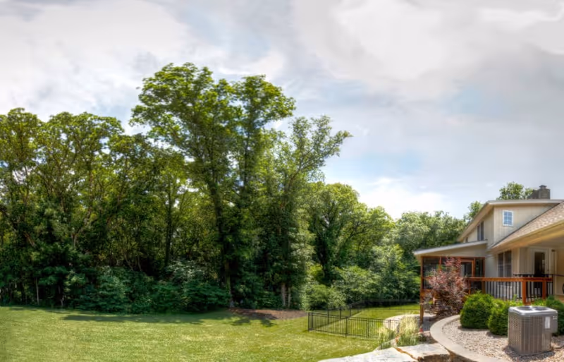 A spacious backyard area of a retirement community with a well-maintained lawn, surrounded by tall green trees. On the right side, part of a building with a covered porch and railing is visible, along with some landscaping and an outdoor air conditioning unit.