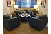 A cozy living room area with two dark blue armchairs facing a round coffee table. Behind the table are two matching sofas with decorative pillows. The room has a window with blinds and two table lamps on side tables next to the sofas. The floor is carpeted in a neutral tone.