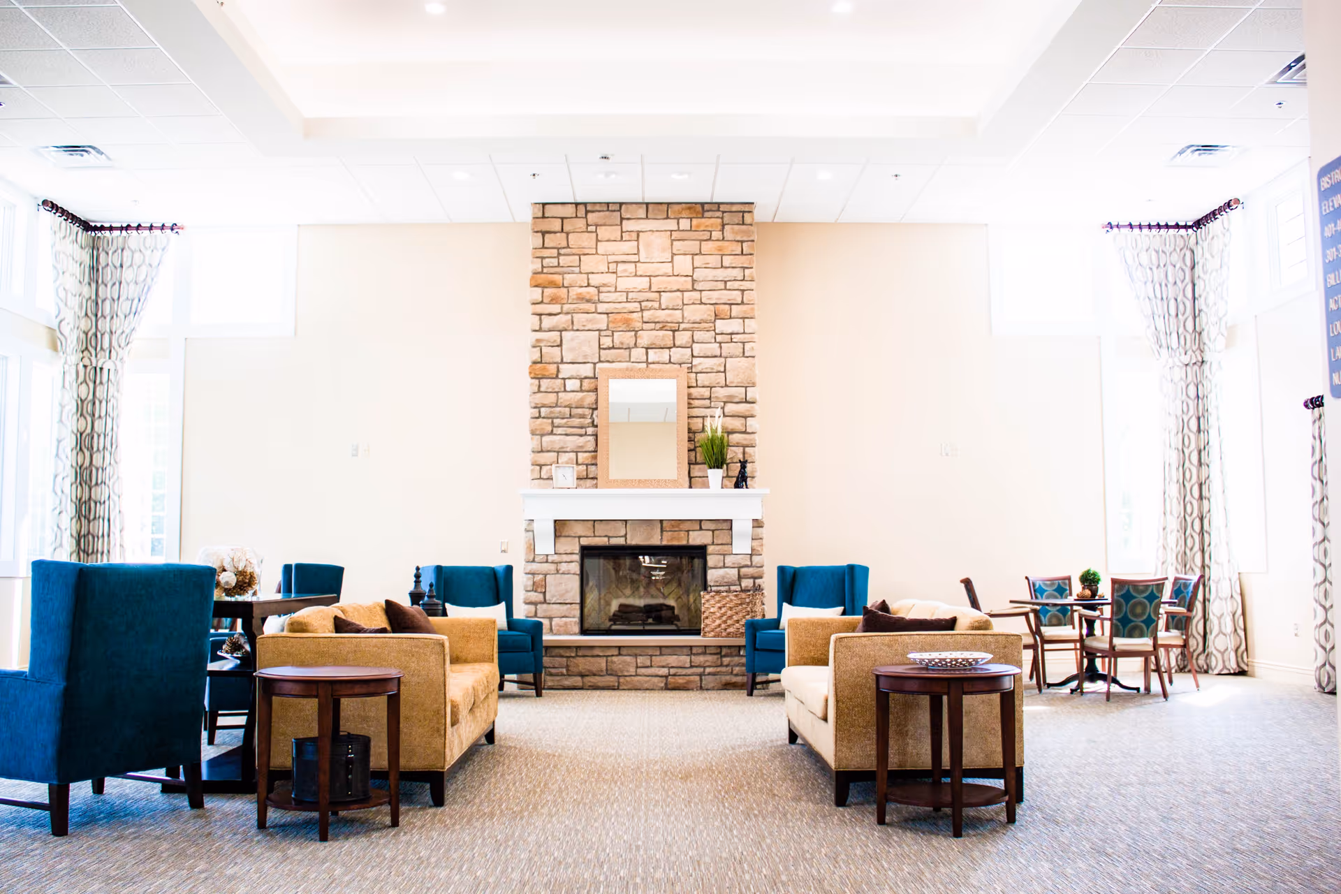 A bright and spacious living room area with a stone fireplace centered on the far wall. The room features beige walls and carpet, two beige sofas with brown pillows facing each other, and four blue armchairs arranged around small wooden tables. Large windows with patterned curtains allow natural light to fill the space. There are also small dining tables with chairs in the background.