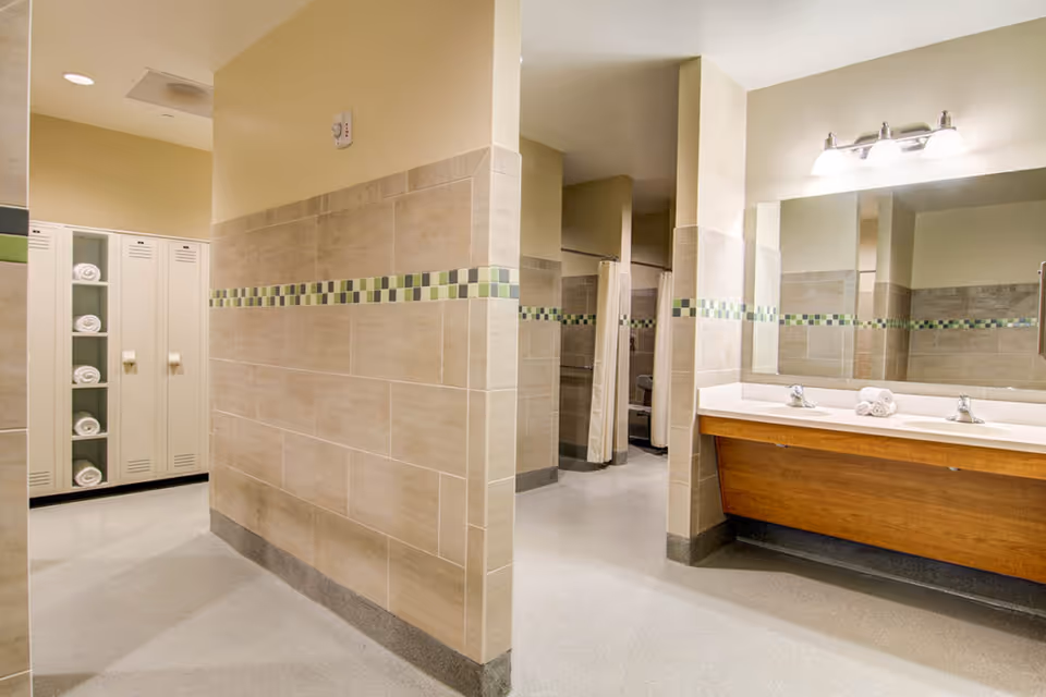 A clean and modern locker room and bathroom area with beige tiled walls featuring a green and gray checkered accent strip. There are white lockers with rolled white towels inside, a large mirror above a double sink with wooden cabinetry, and shower stalls with beige curtains.