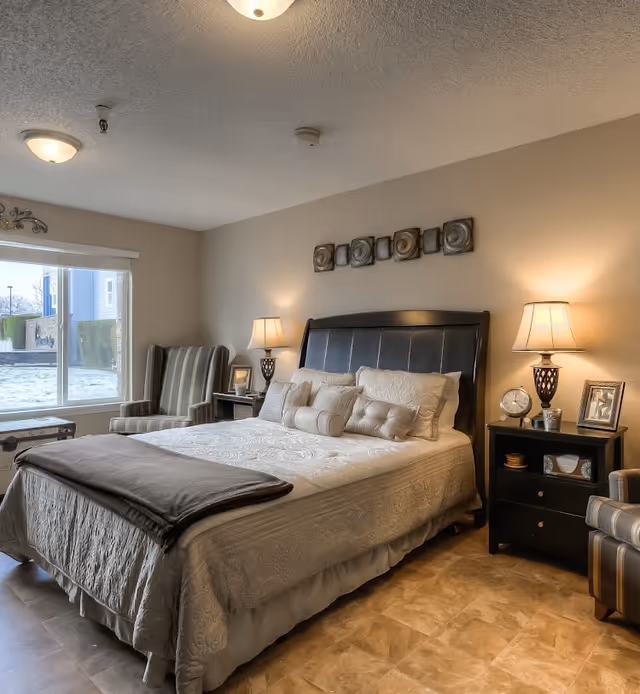 A cozy bedroom with a large bed featuring multiple pillows and a folded blanket at the foot. Two bedside tables with lamps, a clock, and framed photos flank the bed. There are two striped armchairs near a large window that lets in natural light. The walls are painted beige and decorated with a metal wall art piece above the bed.