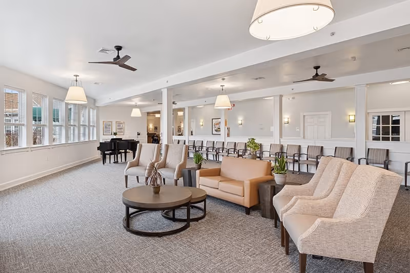 A spacious and well-lit common area with beige armchairs and a sofa arranged around two round coffee tables. The room has large windows on one side, ceiling fans, hanging light fixtures, and a black grand piano in the corner. Additional seating is lined up against the far wall.