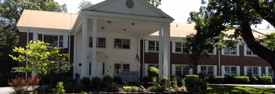 Front exterior view of The Ballou Home, a two-story brick building with white columns supporting a portico entrance, surrounded by trees and landscaped greenery.