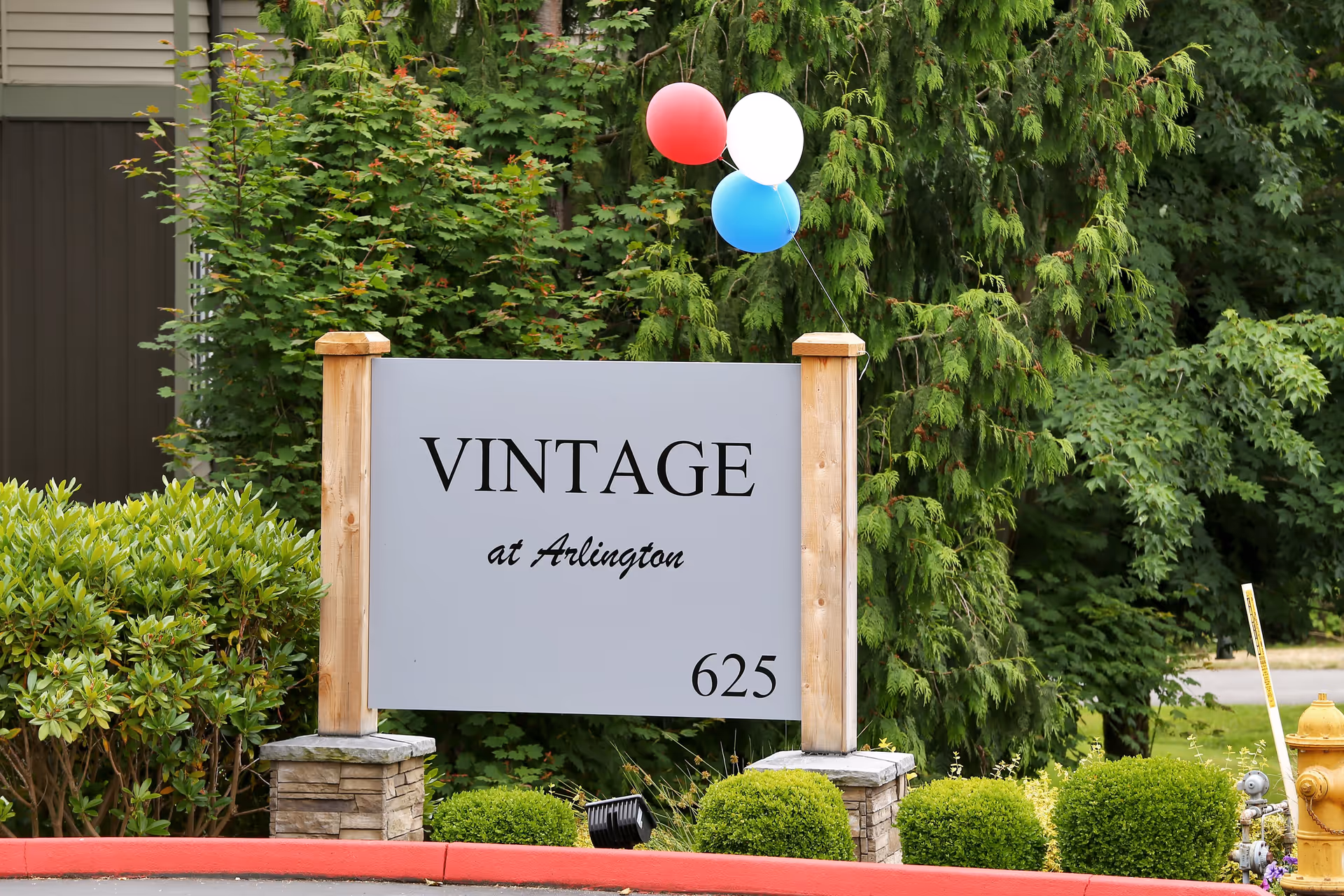 A sign for Vintage at Arlington Senior Apartments with the number 625, surrounded by green bushes and trees. Three balloons in red, white, and blue are tied to the sign.