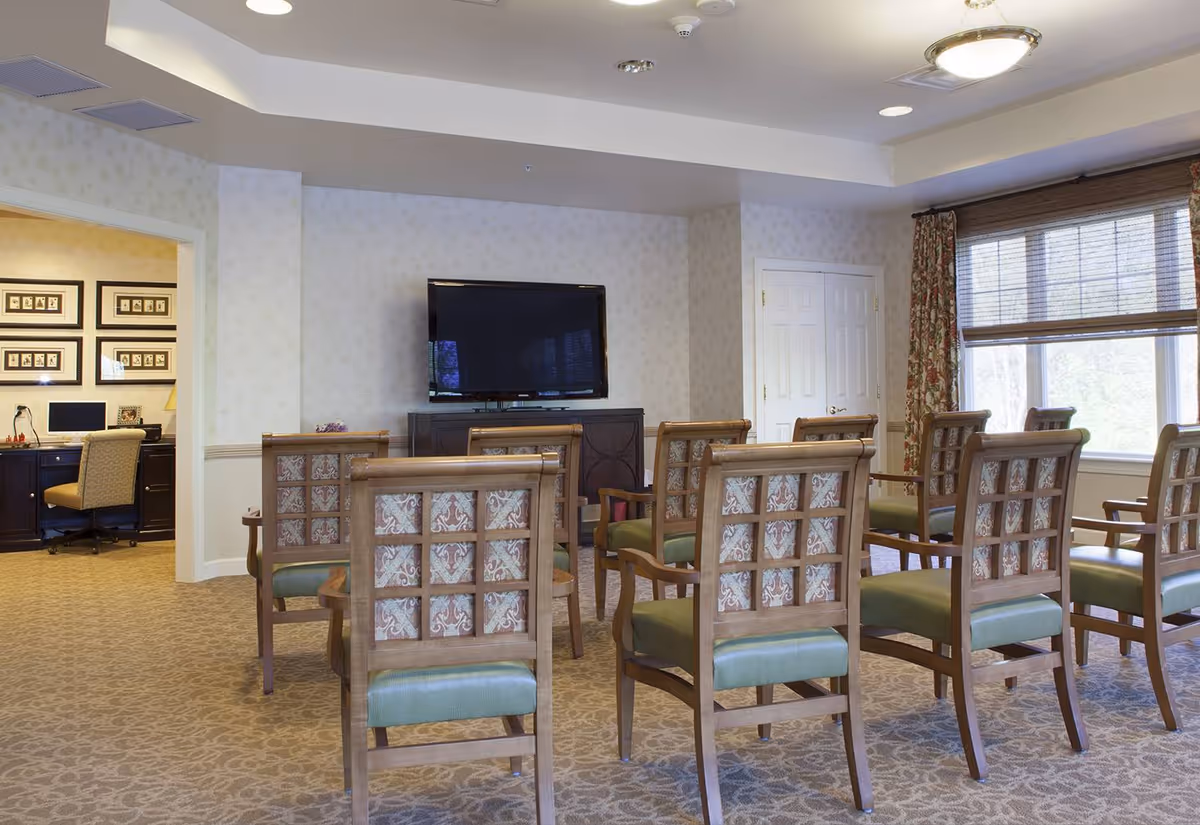 A well-lit common area with multiple wooden chairs arranged in rows facing a flat-screen TV mounted on a cabinet. The room has patterned carpet, floral curtains, and a large window with blinds. An adjacent office space with a desk, chair, and framed pictures on the wall is visible through an open doorway.