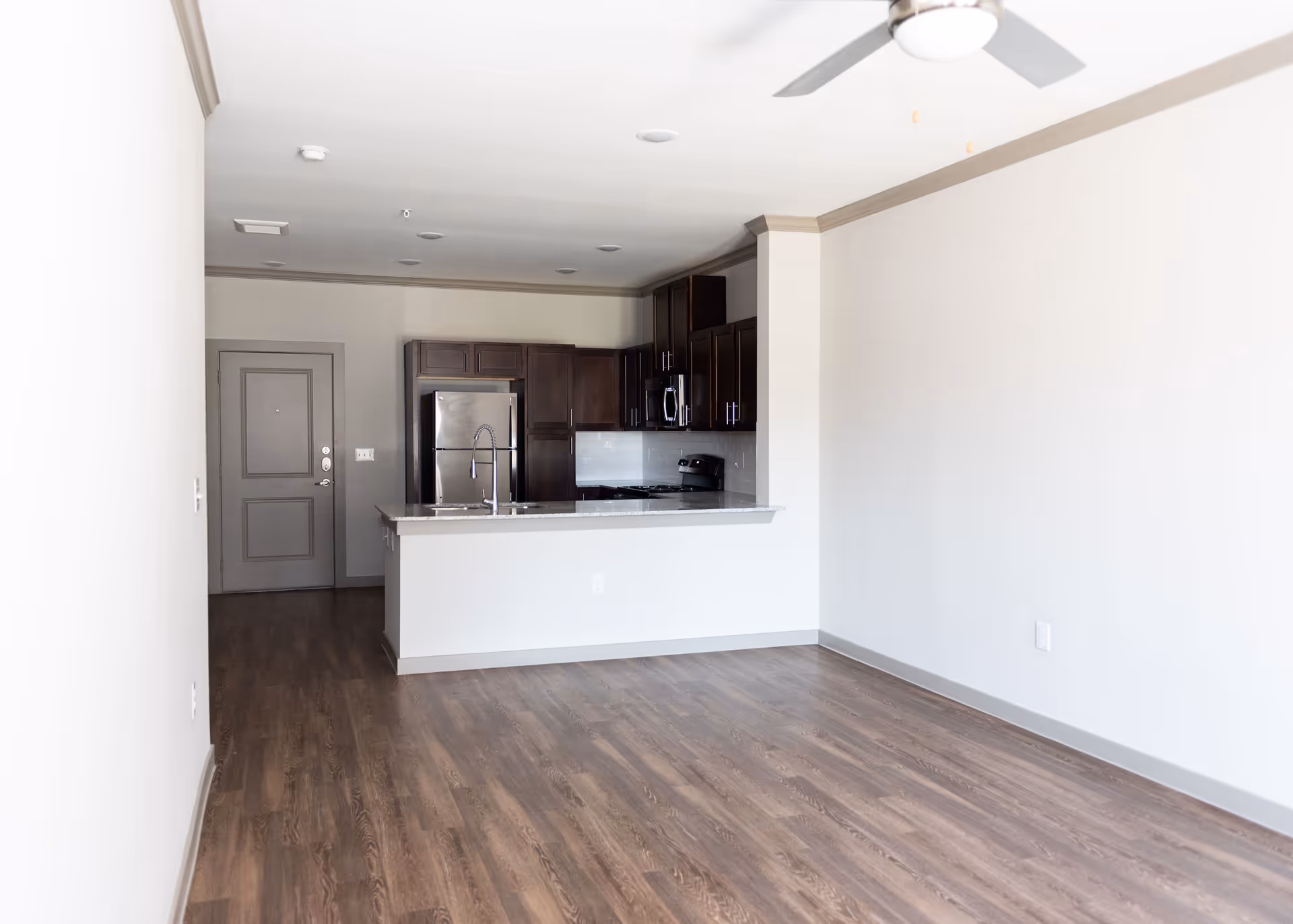 Empty open-plan living area with wood floors, a ceiling fan, and a kitchen with stainless steel appliances and an island.