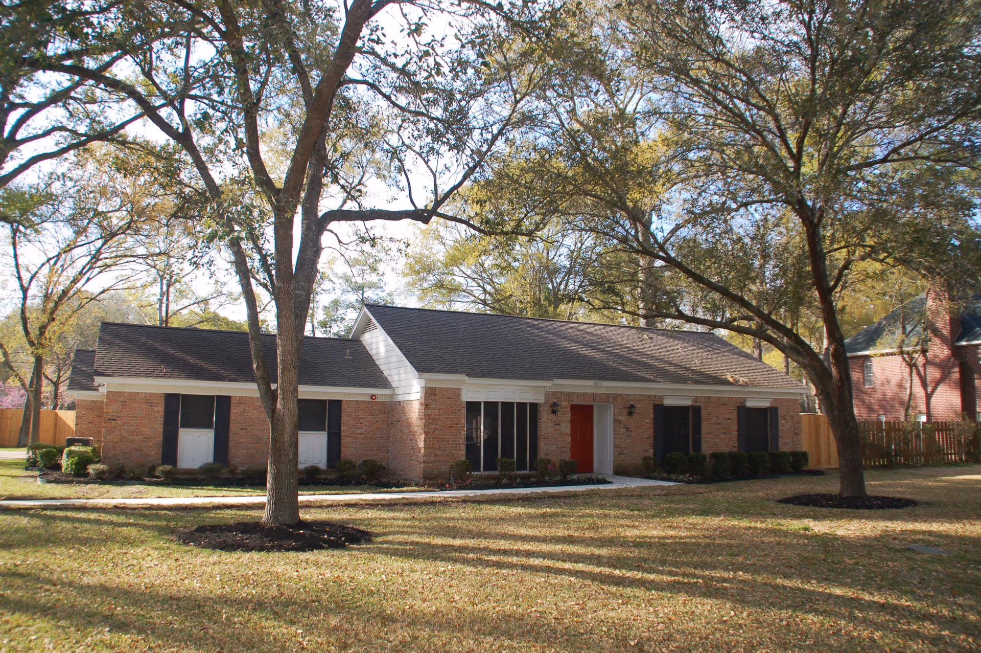 Single-story brick building with a dark shingled roof, several windows with black shutters, and a red front door. The building is surrounded by a grassy yard with large trees casting shadows on the ground. A wooden fence is visible in the background.